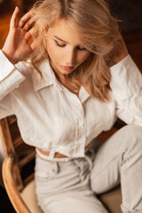 Fototapeta premium Fashion cute blonde girl with beautiful face in trendy elegant white shirt with jeans sits in chair indoors