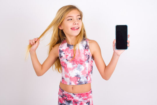 Photo of nice pretty little caucasian kid girl wearing sport clothing over white background demonstrate phone screen hold hair tails