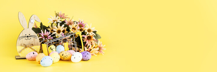 Colorful easter quail eggs in shopping cart, wooden bunny and spring chrysanthemum flowers on yellow background. Copy space. Minimal Easter concept. Banner