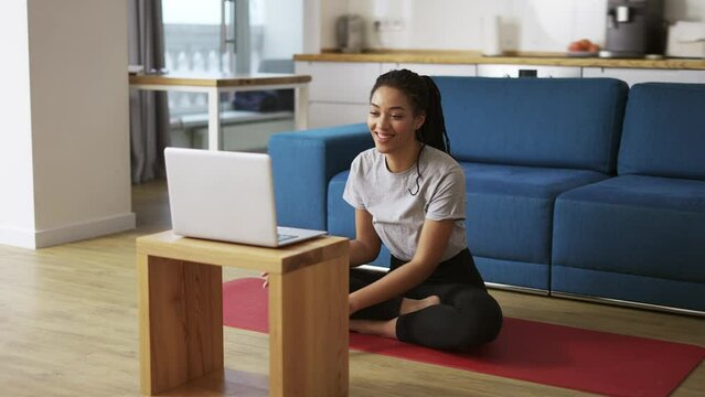 Smiling african american woman talking on video call on laptop while sitting on yoga mat