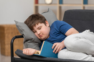 A boy falls asleep while reading a book on the sofa in his room.