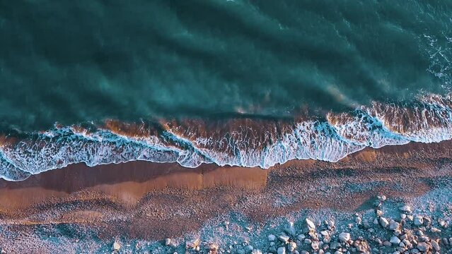 Plano cenital de oleaje en la playa del mar mediterr&aacute;neo con tonos de aguamarina verde y azul espuma y arena marr&oacute;n con piedras