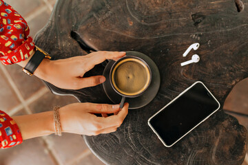 Top view of woman's hands touching a cup of coffee o n the wood background with smartphone and...