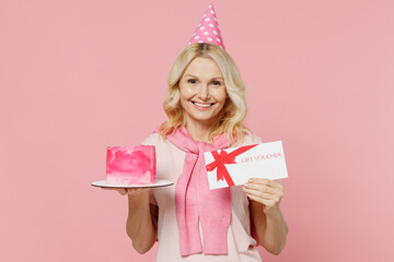 Elderly happy woman 50s wear t-shirt birthday hat hold cake gift certificate coupon voucher card for store isolated on plain pastel pink background studio portrait Celebration party holiday concept.