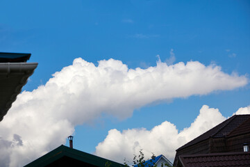 Huge white cloud, in the blue sky, between the roof and chimneys of houses. Beautiful cloudy sky with colorful roofs of houses and tree foliage.