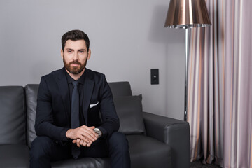 bearded businessman in suit sitting on leather couch in hotel room