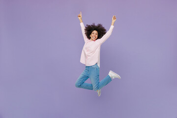 Full body exultant cool jubilant happy excited fun young woman of African American ethnicity wear pink striped shirt jump high with outstretched hands isolated on plain pastel light purple background.
