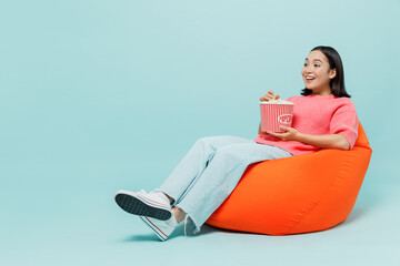 Full body young smiling happy woman of Asian ethnicity 20s wear pink sweater sit in bag chair hold popcorn bucket watch movie film at home isolated on pastel plain light blue color background studio.
