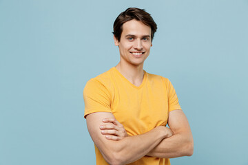 Young smilig happy cheerful confident man 20s wearing yellow t-shirt hold hands crossed folded looking camera isolated on plain pastel light blue background studio portrait. People lifestyle concept.