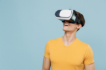 Young amazed surprised caucasian smiling happy fun man 20s in yellow t-shirt watching in vr headset pc gadget isolated on plain pastel light blue background studio portrait. People lifestyle concept