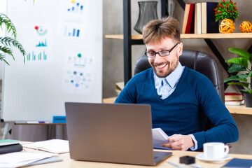 Smiling young confident successful businessman in eyeglasses, formal wear working on online project on laptop in modern office, communicating distantly with client, typing email, do electronic report