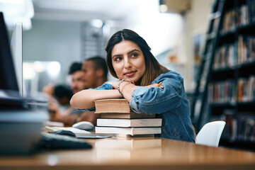 Im going to make you so proud. Shot of a young woman resting on a pile of books in a college library and looking thoughtful. - obrazy, fototapety, plakaty
