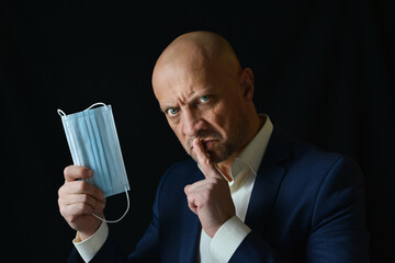 A man in a business suit holds a medical mask in his hand, black background. Demonstration of personal protective equipment against viral infections.