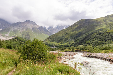 Fototapeta na wymiar Patara Enguri River flowing down the a valley in the Greater Caucasus Mountain Range in Georgia, Svaneti Region, Ushguli. The Shkhara Mountain is covered by clouds. Snow-capped mountains.Glacier