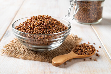 Ingredient for cooking buckwheat porridge concept. Raw organic buckwheat in a glass bowl and wooden spoon on a white wooden tabletop. Gluten free diet and healthy vegetarian food.