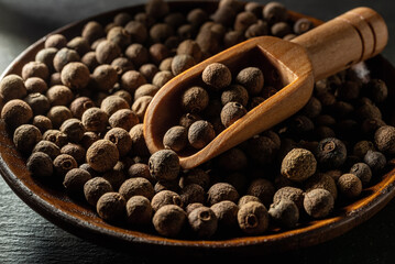 Allspice jamaica pepper in a wooden scoop over allspice grains on a wooden plate. Macro of whole dried pimento berries in the bright sunlight. Organic spices and seasonings concepts.