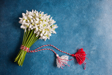 A bouquet of snowdrops flowers and a red and white rope with tassels, a symbol of the arrival of spring on a blue background. Postcard for the feast of March 1 Martisor and Baba Marta, space for text.