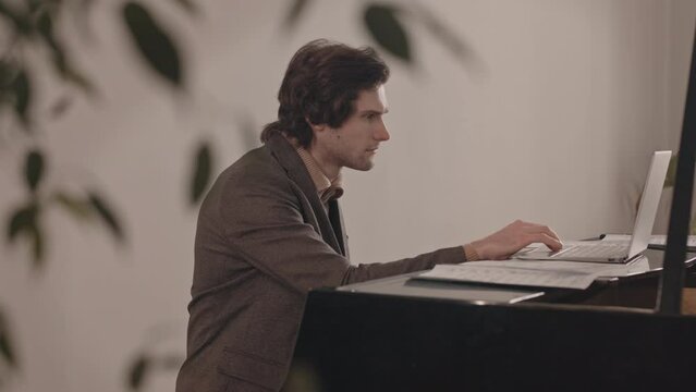 Medium slowmo of young Caucasian man giving online video music lesson on laptop sitting by grand piano indoors
