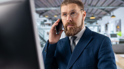 Serious bearded businessman wearing eyeglasses and suit having mobile conversation while working at pc at office. Concept of people, technology and business.