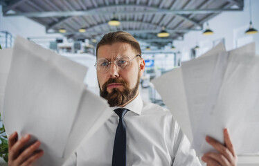 Fototapeta premium Caucasian office worker with stack of paper in hands feeling frustration from overwork at office. Bearded man in eyewear and white shirt having emotional stress.
