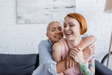Fototapeta na wymiar joyful lesbian woman with closed eyes embracing tattooed girlfriend at home