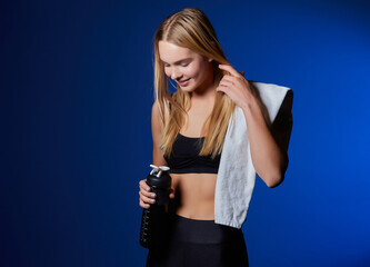 Sports girl, with a towel on her shoulder and a bottle of water in her hands, is resting after training in the gym