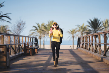Woman listening to music with headphones and mobile phone while walking on a bridge at the coast