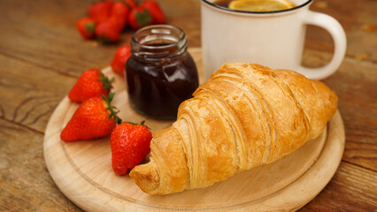 Tea break snack. Lemon tea, croissant, jar of jam and fresh strawberries on wooden background. Sweet tasty breakfast. Focus on freshly baked croissant