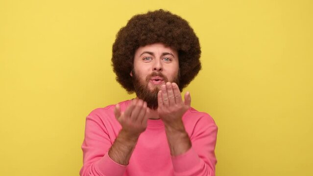 Happy bearded man with Afro hairstyle looking at camera and sending air kiss, expressing romantic feelings, wearing pink sweatshirt. Indoor studio shot isolated on yellow background.
