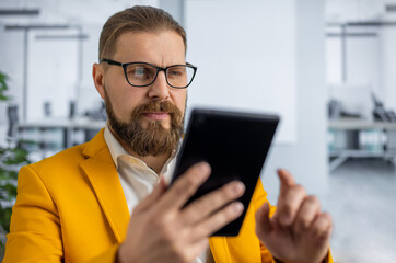 Handsome bearded man in bright yellow jacket and eyewear using digital tablet for work at office. Concept of people, confidence and gadgets.