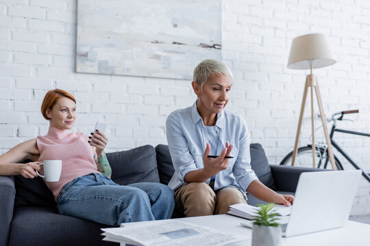 lesbian woman working on laptop near girlfriend sitting on sofa with smartphone and cup of tea - Powered by Adobe