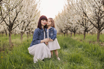 Portrait of beautiful young mother posing in embrace with her little cute daughter among blooming apple garden. Stylish family look of white delicate dresses and denim jackets.