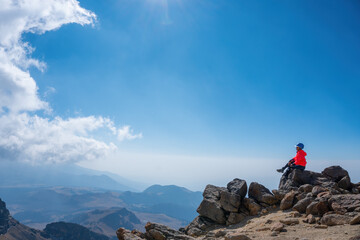 Fototapeta premium A young cheerful girl in a helmet, sits on the top of the mountain higher than clouds
