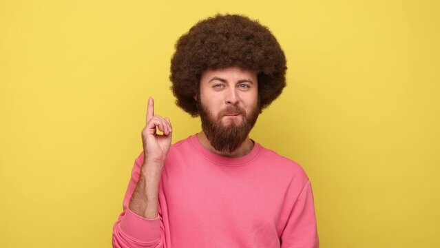 Bearded man with Afro hairstyle raising finger having genius idea, looking amazed by sudden smart solution, creative thought, wearing pink sweatshirt. Indoor studio shot isolated on yellow background.