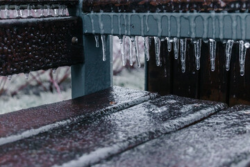 A wooden brown bench covers a layer of ice and icicles after freezing rain in winter