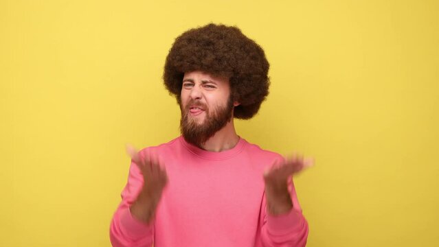 Bearded angry man with Afro hairstyle has problems, gesticulating with hands, crisis and negativity, misunderstanding, wearing pink sweatshirt. Indoor studio shot isolated on yellow background.