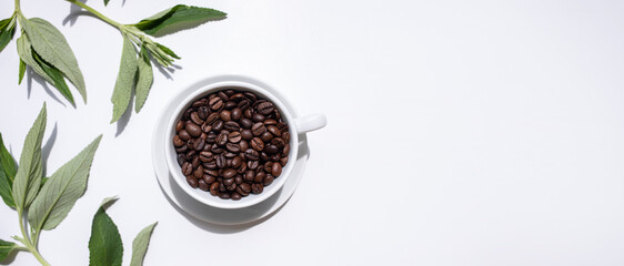 Coffee beans in a white cup on a white background among green coffee leaves. Banner, top view, place for text.