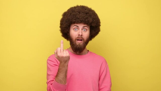 Bearded man with Afro hairstyle scratching his head and showing middle finger to camera with amazed facial expression, wearing pink sweatshirt. Indoor studio shot isolated on yellow background.
