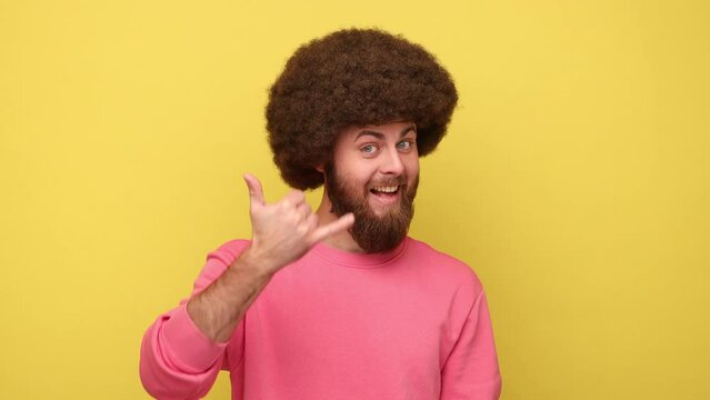 Bearded man with Afro hairstyle standing with telephone hand gesture smiling to camera, flirting offering to contact by phone, wearing pink sweatshirt. Indoor studio shot isolated on yellow background