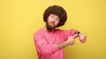 Bearded man with Afro hairstyle pointing finger at wrist watch and looking annoyed and displeased, showing clock to hurry up, wearing pink sweatshirt. Indoor studio shot isolated on yellow background. - Powered by Adobe