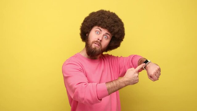 Bearded man with Afro hairstyle pointing finger at wrist watch and looking annoyed and displeased, showing clock to hurry up, wearing pink sweatshirt. Indoor studio shot isolated on yellow background.
