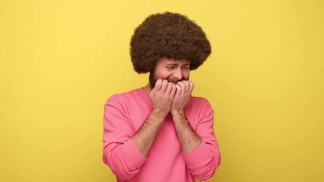 Bearded man with Afro hairstyle biting nails on fingers looking around with terrified expression, confused worried about work, wearing pink sweatshirt. Indoor studio shot isolated on yellow background