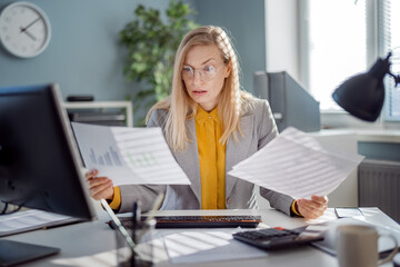 Mature caucasian lady in eyeglasses and formal suit examining various documents while sitting at...
