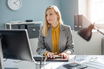 Caucasian female manager with blond hair sitting at office desk and typing on keyboard of modern...