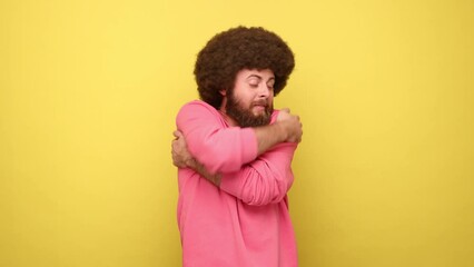 Bearded smiling man with Afro hairstyle hugging himself, egoistic proud of his achievements, encouraging, wearing pink sweatshirt. Indoor studio shot isolated on yellow background.