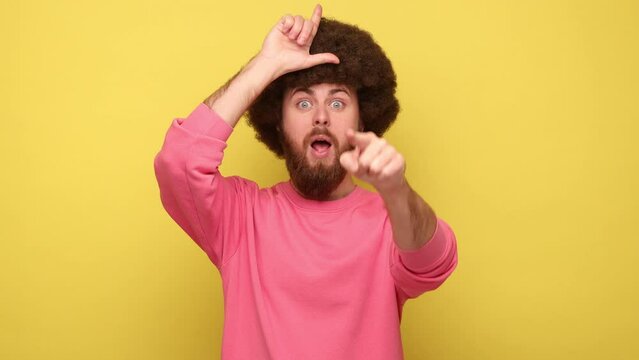You are looser. Bearded man with Afro hairstyle showing loser gesture and pointing at camera, mocking your defeat, wearing pink sweatshirt. Indoor studio shot isolated on yellow background.