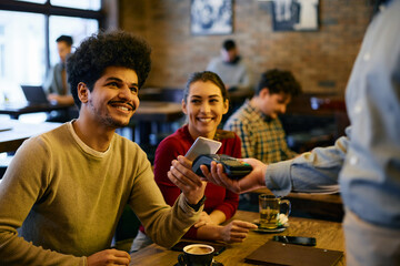 Young man making contactless payment with smart phone while being with his girlfriend in a cafe.
