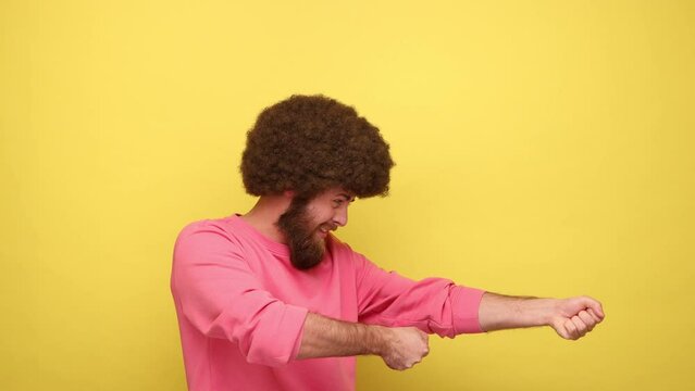Side view of bearded man with Afro hairstyle pretending to hold rope in hands and pull, expressing much effort on face, wearing pink sweatshirt. Indoor studio shot isolated on yellow background.