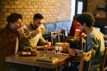 Multiracial group of male friends drink beer while having lunch in pub.