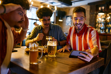 Young men drink beer while watching sports match on smart phone in pub.